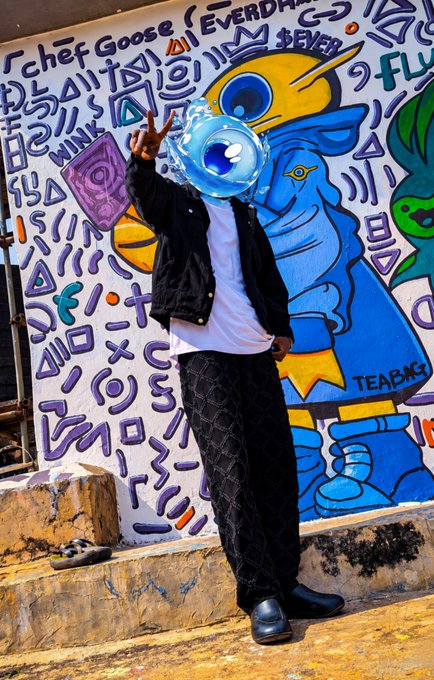 A person poses in a black jacket and patterned pants in front of a colorful graffiti wall featuring a blue character, making a peace sign with their face obscured by a digital sphere.