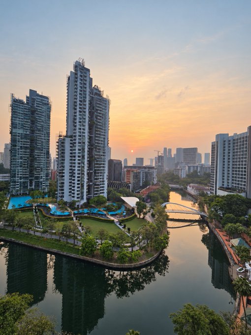An aerial view of a modern city at sunset, featuring tall residential skyscrapers, a swimming pool area, green parkland, and a river with arched bridges, all bathed in warm golden light.