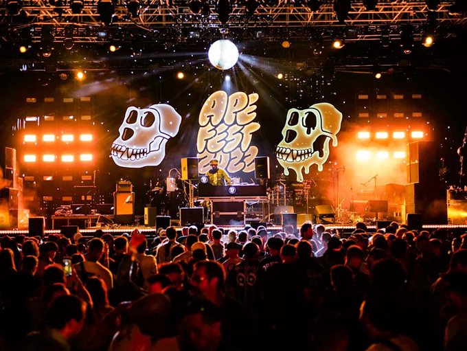A large crowd gathers at an "APE FEST '22" concert, with two glowing Bored Ape skull designs flanking the stage beneath a disco ball, directly illustrating the post's reference to a Bored Ape NFT rave celebration.