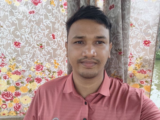 A selfie of a young man with dark hair and a light beard wearing a pink/coral button-up shirt, posed in front of floral patterned curtains.