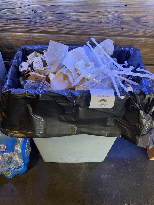 An overflowing trash can filled with discarded paper receipts, plastic utensils, and wrappers sits next to a pack of bottled water, with one receipt partially visible reading "SECONDARY MARKET" — humorously implying that people are literally "throwing away" their Bitcoin receipts.