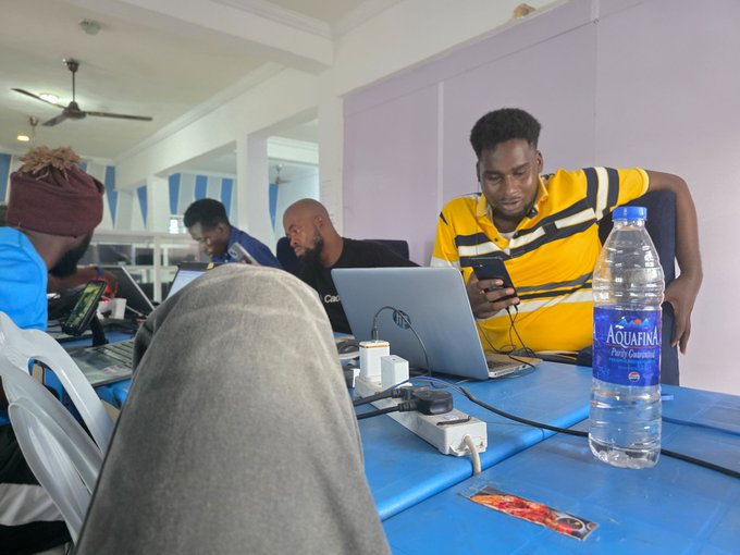 Several men work on laptops at a blue table in what appears to be a workspace or internet cafe, with one man in a yellow and black striped shirt using a phone next to a bottle of Aquafina water.