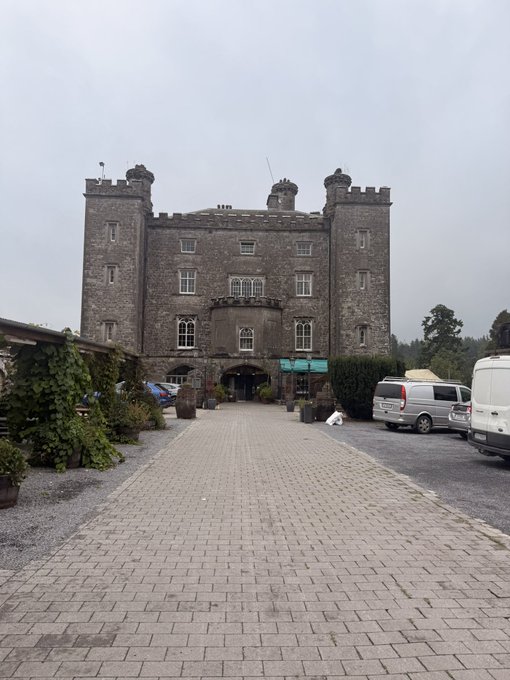 A photograph of a large, historic stone building featuring castle-like turrets and battlements, with a stone-paved driveway leading to a central arched entrance, parked vans on the side, and a bag of supplies sitting on the ground.