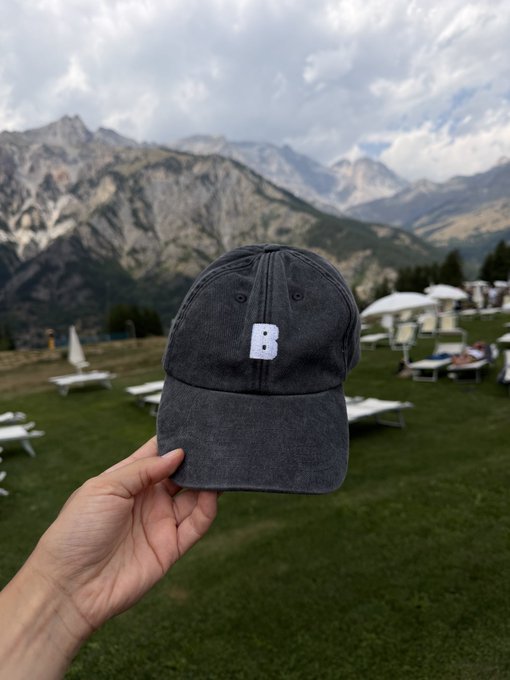 A hand holding a faded black baseball cap with a white letter "B" embroidered on it, with a scenic view of mountains and lounge chairs in the background.