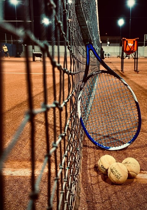 A blue tennis racket leaning against a chain-link net on a clay court at night, with three tennis balls at its base and stadium lights glowing in the background.