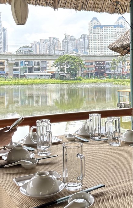A view from a thatched-roof restaurant overlooking a river with urban buildings and an elevated highway in the background, with a table set with bowls, chopsticks, and glasses ready for dining.