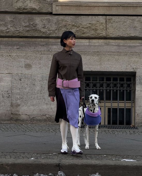 A woman wearing a brown shirt, purple skirt, white tights, and a pink belt stands next to a Dalmatian dog wearing a blue sweater against a stone wall.