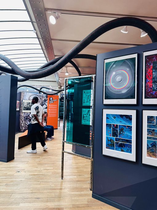 An art gallery or exhibition space displays framed artworks on dark blue walls, with visitors browsing near large black ventilation tubes overhead.