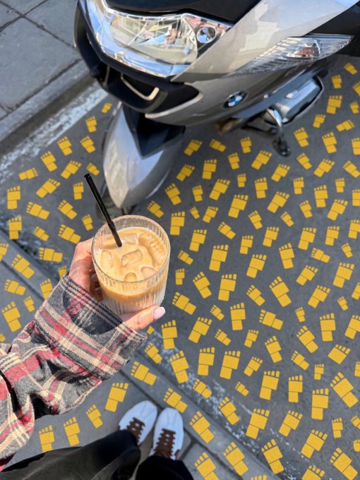 A first-person view of a hand holding an iced coffee drink over a sidewalk decorated with yellow footprints pointing toward a parked scooter.