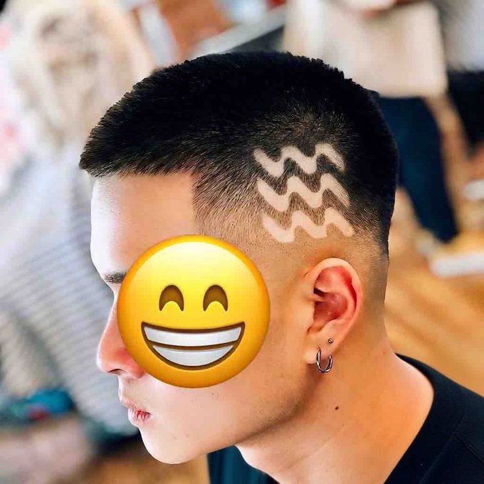 Photograph of a person with a short buzz-cut hairstyle featuring the Aquarius zodiac symbol (wavy lines) shaved into the side, with a smiling emoji covering their face.