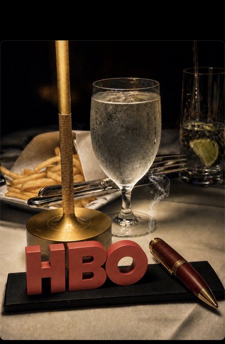 A promotional image featuring the red 'HBO' logo in 3D letters on a dark surface, alongside a brass gavel, a glass of water, a wine glass with a lime, french fries, and a brown pen.