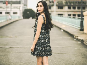A young woman with long dark hair stands on a bridge wearing a sleeveless black lace dress, looking back over her shoulder toward the camera.