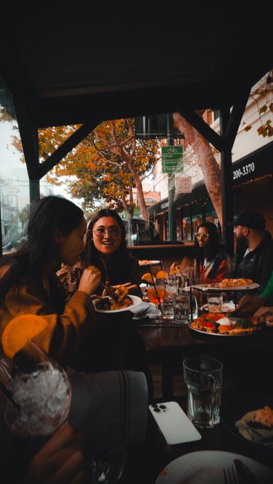 A group of friends enjoys a lively meal at an outdoor restaurant, with plates of food and drinks scattered across the table as autumn trees and a storefront numbered "206-3370" frame the scene. The image captures the casual, real-world dining experience that the post references, illustrating the everyday local restaurants that form a network of over 2,500 merchants for builders to connect with.