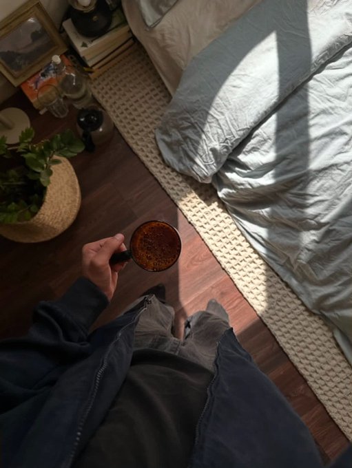 A first-person view shows a hand holding a mug of coffee while looking down at a bedroom floor with a bed, rug, potted plant, and bedside table.