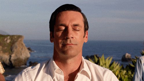 The image features an actor posing for a photo with his eyes closed, possibly meditating or taking a break from work. The background of the image shows a beautiful ocean view, adding to the serene atmosphere. There are two other people present in the scene, one on the left side and another person standing further back towards the right edge of the frame.