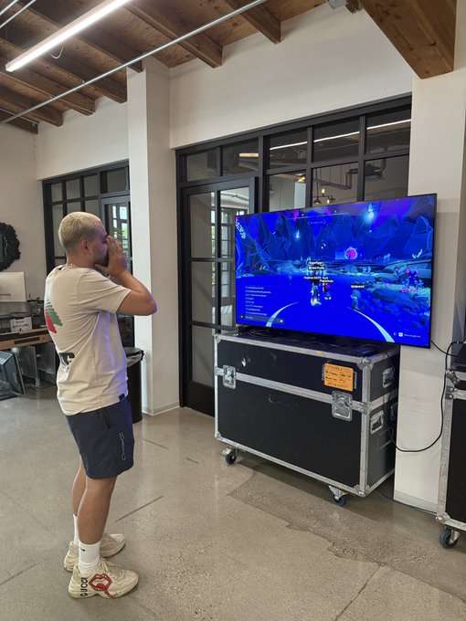 A man in a t-shirt and shorts stands watching a video game being played on a large TV resting on a black road case.