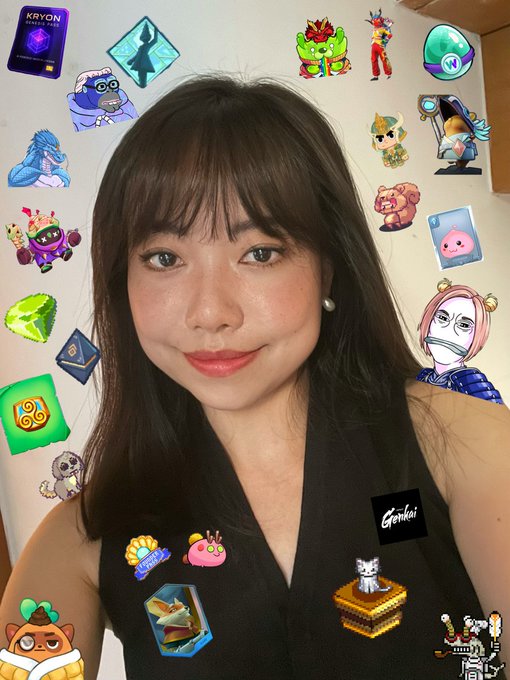 The image features a woman with long brown hair wearing a black shirt, posing for a selfie that includes various stickers and logos from different games or projects. The woman's face is partially obscured by these decorations, making her appear like she is hiding behind them. The background of the photo shows a white wall, which contrasts nicely with the colorful elements on the woman's shirt.