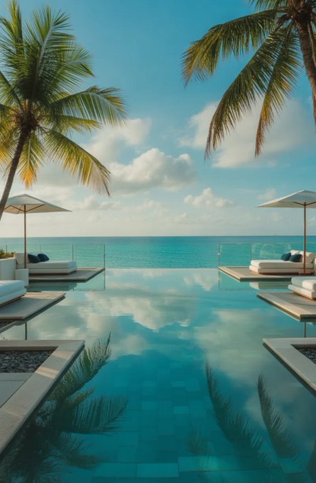 The image shows a large swimming pool with two white lounge chairs, one of which has an umbrella attached. There are also palm trees surrounding the pool area. In the background, there is a view of the ocean or sea, adding to the relaxing atmosphere. The photo appears to be taken from the side of the pool, providing a clear view of the surroundings and the water's edge.