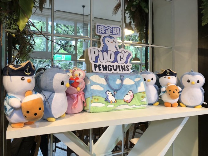 A display of PUDGY PENGUINS merchandise, including various plush penguin toys in different outfits and a tote bag, arranged on a white table with the Pudgy Penguins logo sign in the center.