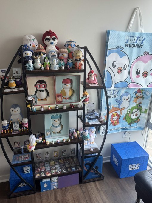 A Pudgy Penguins collection display featuring numerous plush toys, figurines, and accessories arranged on a multi-tiered shelving unit, alongside a "Pudgy Penguins" banner and a blue mystery box on the floor. The display showcases the collector's 6-month journey of acquiring items from Pudgy Penguins mystery boxes, with various sized penguins and merchandise organized throughout the shelves and bottom storage compartments.