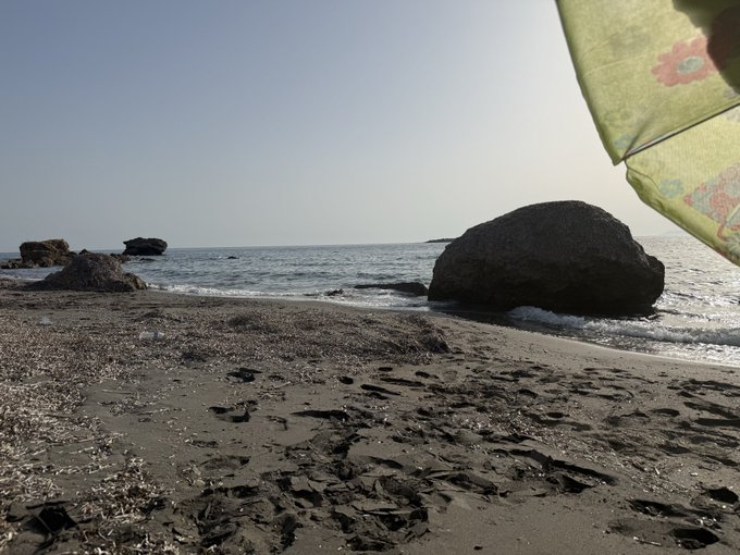 A beach scene showing a sandy shore with footprints, large rocks in the water, and a yellow patterned umbrella partially visible on the right side.