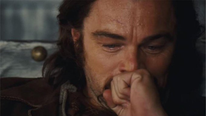 A close-up cinematic shot of a man with long hair, his face dirty and tearful, biting his fist in apparent anguish, with a blue and gold fabric backdrop behind him.
