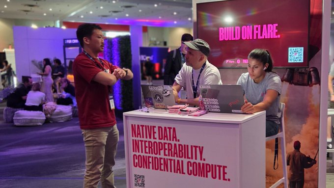 The image shows a group of people gathered around a booth with laptops, possibly attending an event or exhibition related to technology. There are three individuals standing near the booth, while another person can be seen sitting at the table. The booth features a sign that reads "Native data, Interoperability, Confidentiality, Computer".
