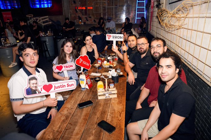 A group of people seated at a long wooden table at a TRON/Justin Sun fan meetup, holding branded signs with hearts and photos.