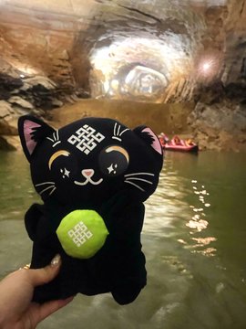 A black plush toy cat holding a green ball is held in the foreground of a cave with an underground lake where people are visible in red boats.