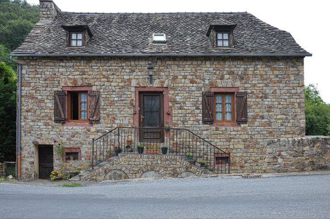 A traditional stone house with a slate roof, dormer windows, and wooden shutters, featuring a staircase entrance with potted plants.