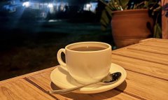 The image features a white cup of coffee sitting on top of a wooden table with a spoon placed inside it. The background shows a night view through a window or patio door, creating an outdoor setting for enjoying a warm beverage. The photo appears to be taken from a low angle, emphasizing the height and prominence of the cup of coffee as the central focus of the scene.