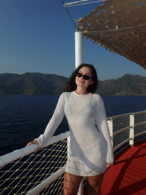 A young woman wearing sunglasses and a white sheer coverup stands smiling on a boat deck with a red carpet, with deep blue water and mountains in the background.