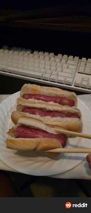 Three sliced hot dogs in buns stacked on a paper plate, held by chopsticks, with a white keyboard in the background.