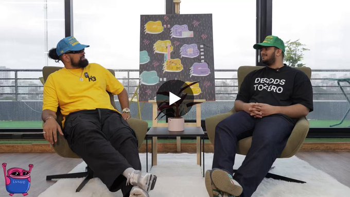 Two men sit in armchairs on a rooftop terrace during what appears to be a video interview, with a play button overlay visible.