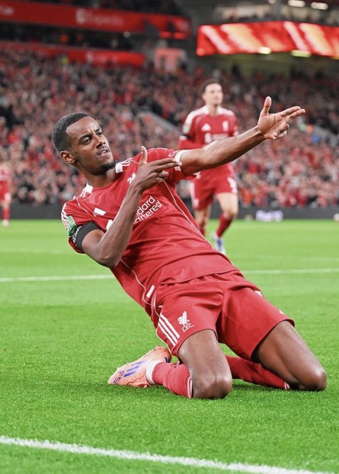 A photograph of a Liverpool soccer player (wearing a red kit with 'standard chartered' on the front) sliding on his knees to celebrate a goal on the pitch.