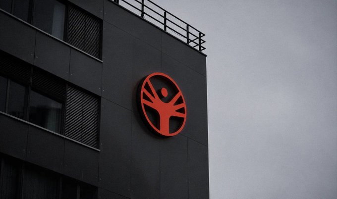 A modern dark gray building features a large red circular logo depicting a stylized human figure with raised arms mounted on its exterior wall.