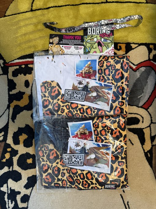 Two leopard-print merchandise items in plastic packaging branded 'BORING' and 'NEVER GONNA BROKE' lie on a patterned blanket, complete with a thank you card.