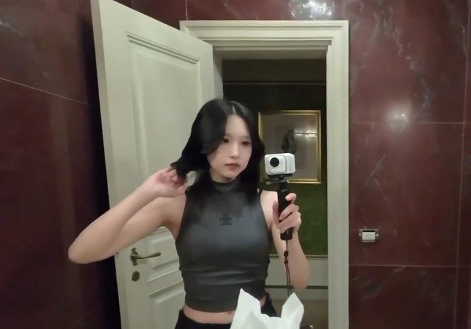 A woman with long dark hair takes a mirror selfie in a marble bathroom, holding a small camera and adjusting her hair.