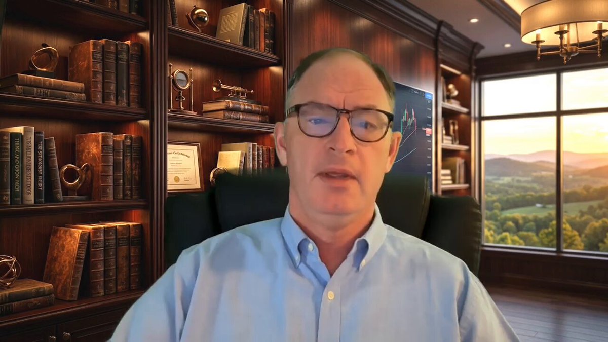 The image shows a man wearing glasses sitting at a desk with bookshelves behind him, suggesting that he might be in an office or a library setting. The man appears to be talking into a microphone, possibly giving a presentation or participating in a video call. There is also a clock on the wall above his head and a window next to it, allowing natural light to enter the room.