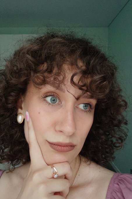 A close-up selfie of a young woman with curly brown hair and blue eyes, wearing pearl earrings and a pink top, posing with her hand on her cheek.