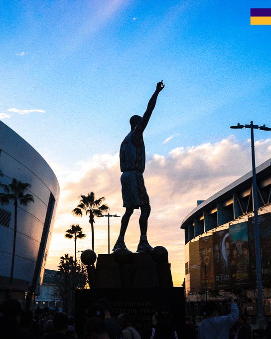 A bronze statue of a basketball player in a triumphant shooting pose stands silhouetted against a sunset sky outside the STAPLES Center in Los Angeles. At the base, a banner reads "SAVE THE GAME," and crowds gather below as palm trees frame the iconic arena.