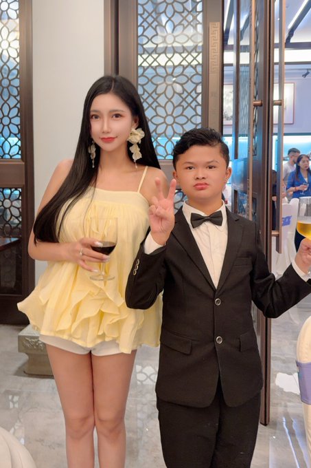 A young woman in a yellow ruffled dress holding a wine glass stands next to a young boy in a black suit with a bowtie; both are making peace signs at the camera.