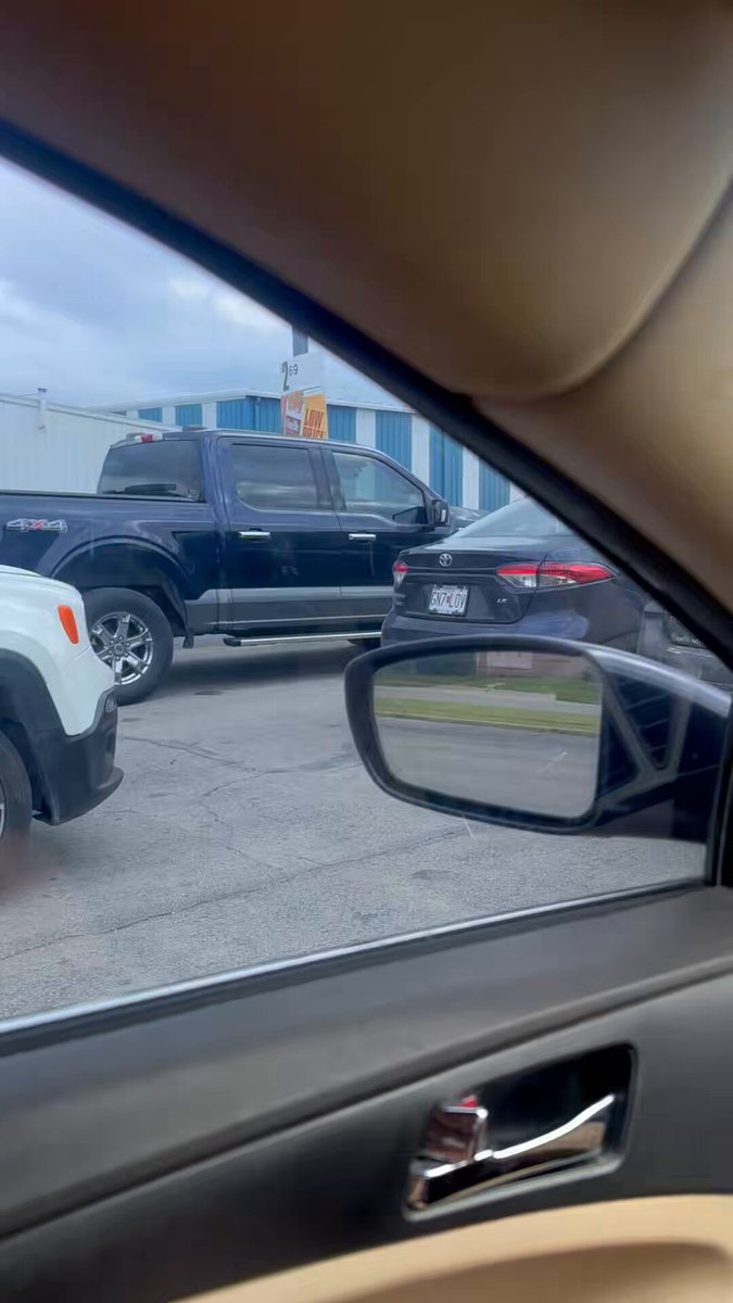 The image shows a car's side view mirror, which has a black dog sticker on it. The mirror reflects the car behind it, including another truck parked nearby. The scene takes place outside of an office building or warehouse, with a parking lot visible in the background.