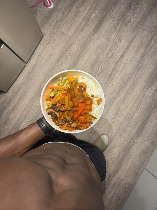 A first-person view looking down at a bowl of rice topped with beef and vegetables, held by a shirtless person standing on a wood-grain floor.