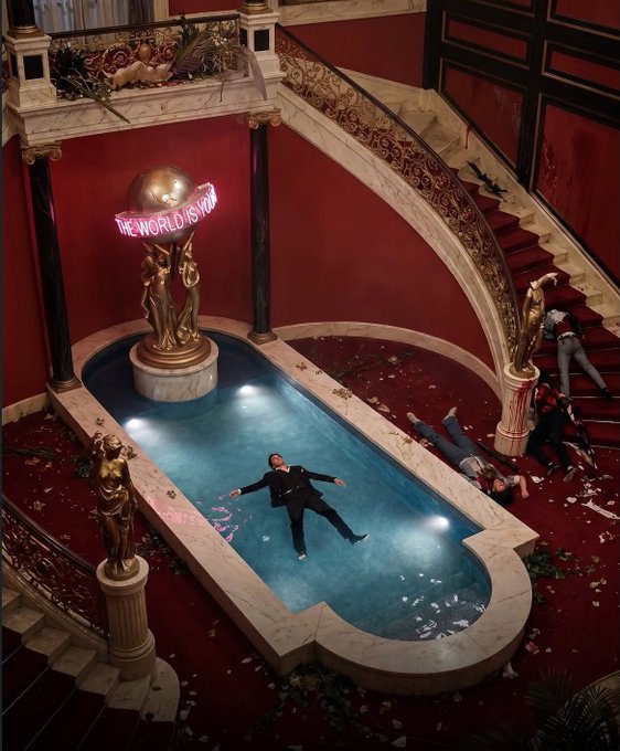 An opulent red and gold mansion interior with a swimming pool, featuring a golden globe statue with neon 'THE WORLD IS YOURS' text, where several people in suits lie motionless on the floor.