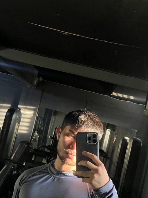 A young man takes a gym mirror selfie with his phone, wearing a light blue long-sleeve athletic shirt, with gym equipment visible in the background.