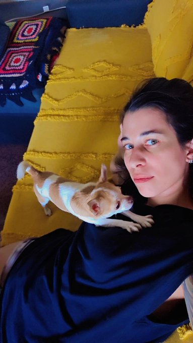 A selfie of a woman lying on a couch with a small white-and-tan Chihuahua dog, with yellow pillows and a colorful crocheted blanket visible in the background.