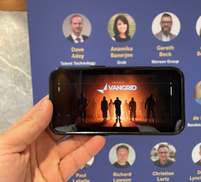 A hand holding a smartphone displaying the 'VANGRID' logo, with a background board showing headshots and names including Dave Adey, Anamika Banerjee, and Gareth Beck.