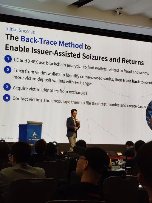A presenter on stage at an AVA Awards event discusses 'The Back-Trace Method to Enable Issuer-Assisted Seizures and Returns,' with a slide outlining four steps including blockchain analytics from LE and XREX to trace funds and contact victims.