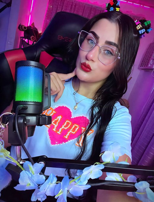 A woman with dark hair, glasses, and a 'HAPPY' t-shirt posing in front of a gaming setup with a colorful RGB microphone and blue flower accessories.